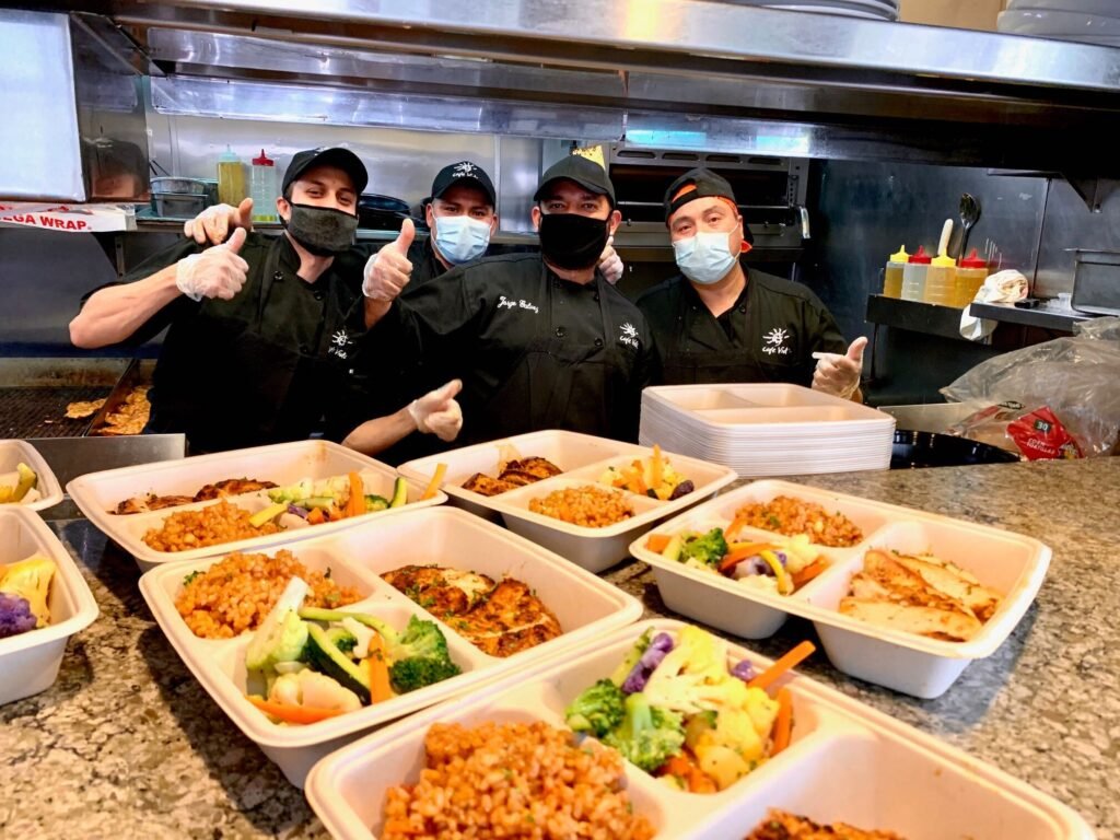 Photo of some of the Cafe Vida kitchen team. The team is wearing gloves and face-masks, and giving thumbs up to the camera. To go orders are shown in the foreground.